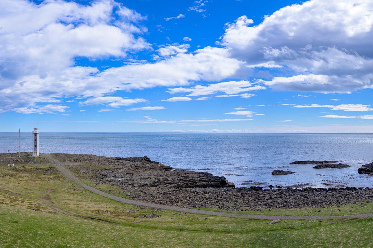 A panoramic view places a slender white lighthouse between a green headland, black lava shore, and the open Atlantic.
