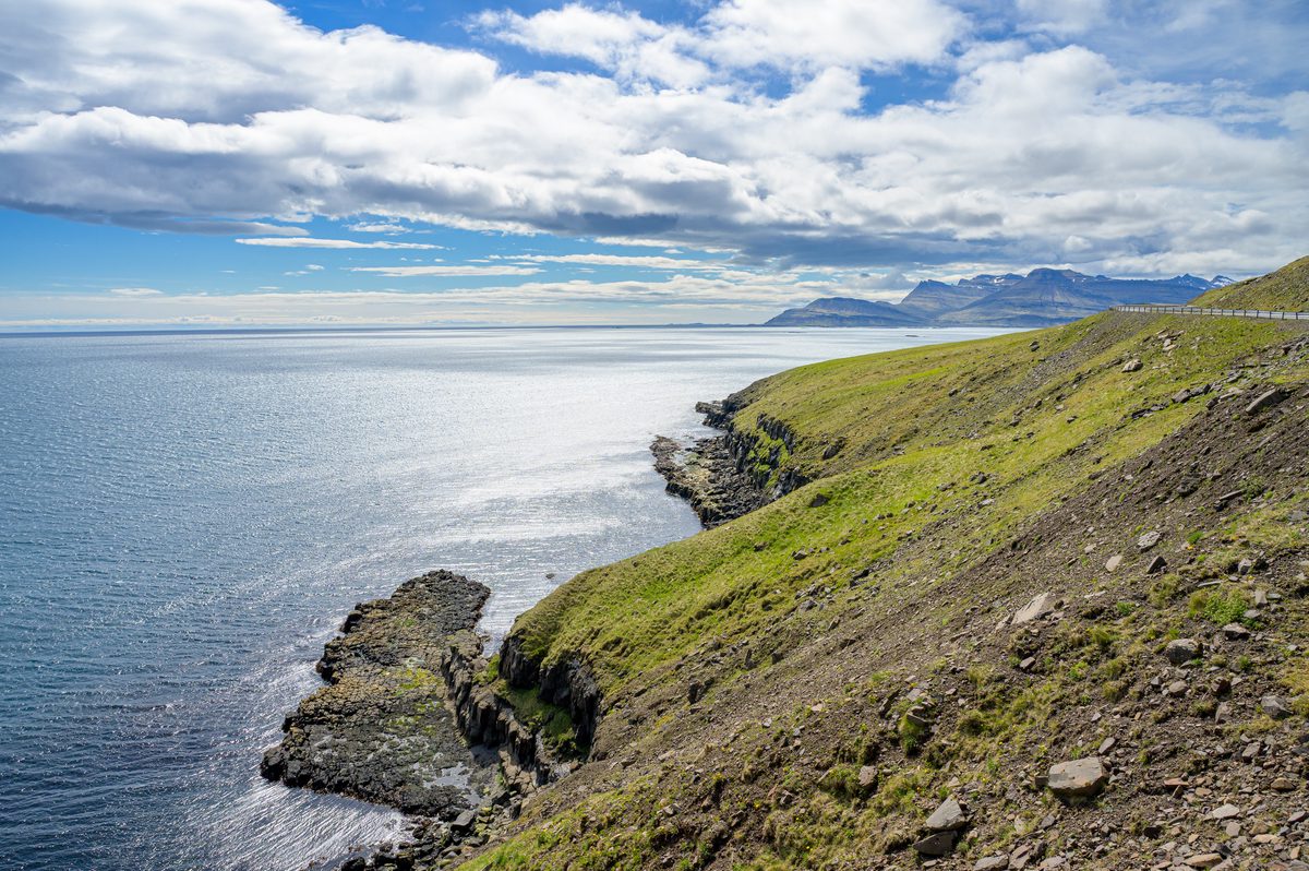 Bright light spreads across the sea below a narrow road carved into the green coastal slope.