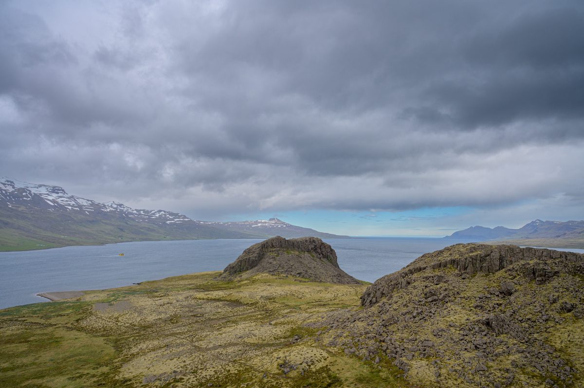Dark weather closes over a broad fjord framed by low volcanic outcrops and snow-patched mountains.