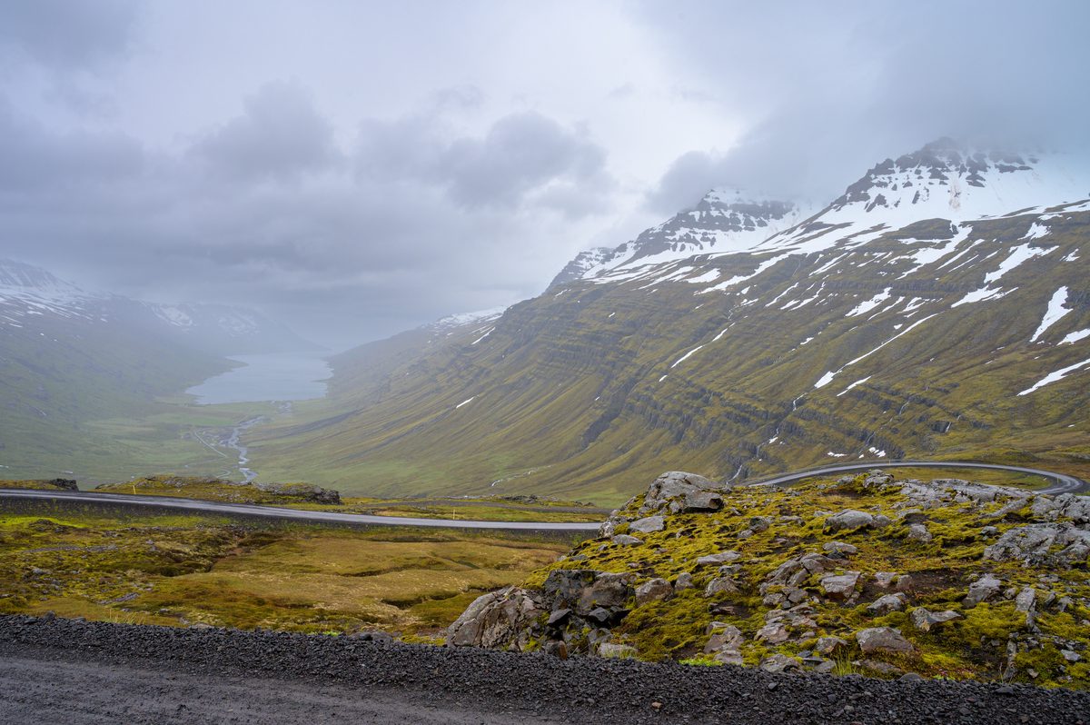 A switchback road drops from the misty pass toward the narrow fjord between steep snow-streaked slopes.