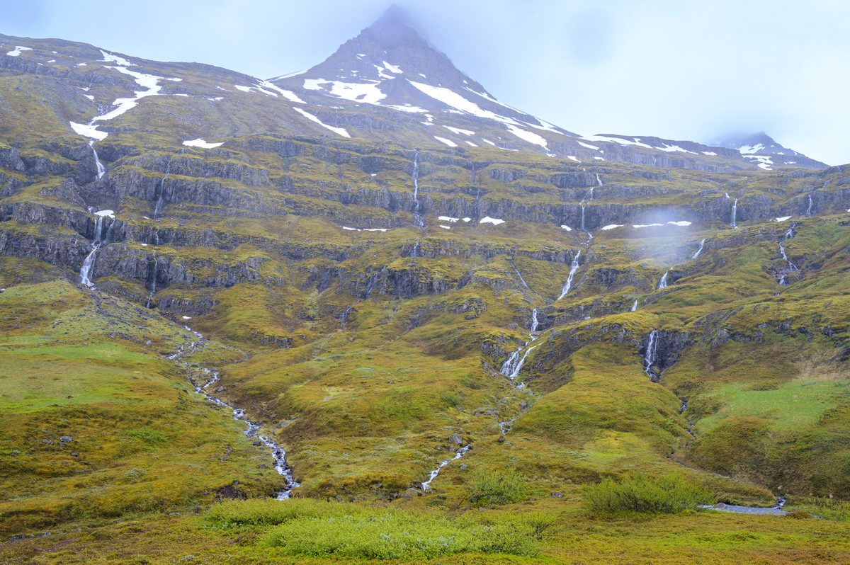Many slender streams of Klifbrekkufossar descend the green terraced mountainside below a cloud-wrapped peak.