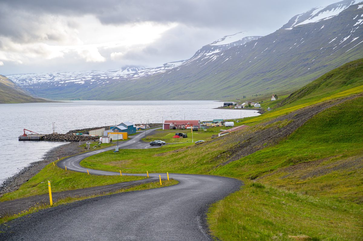 A ribbon of road descends past harbor buildings toward the tiny settlement on the green shore of Mjóifjörður.