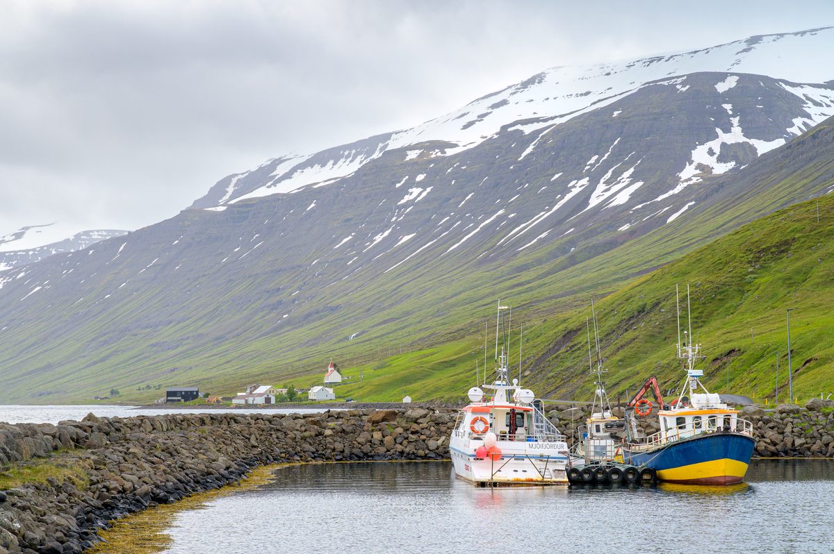 Two working boats rest inside the small stone harbor with the fjord church and mountains beyond.