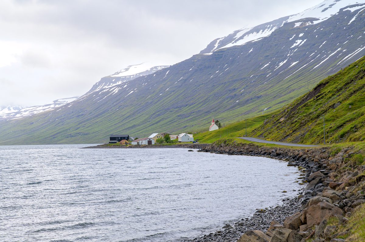 The narrow road follows Mjóifjörður toward a cluster of houses and the small red-roofed church.