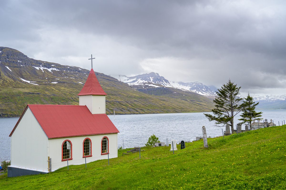A small white church and its graveyard sit on bright grass above the water beneath rain-heavy mountains.