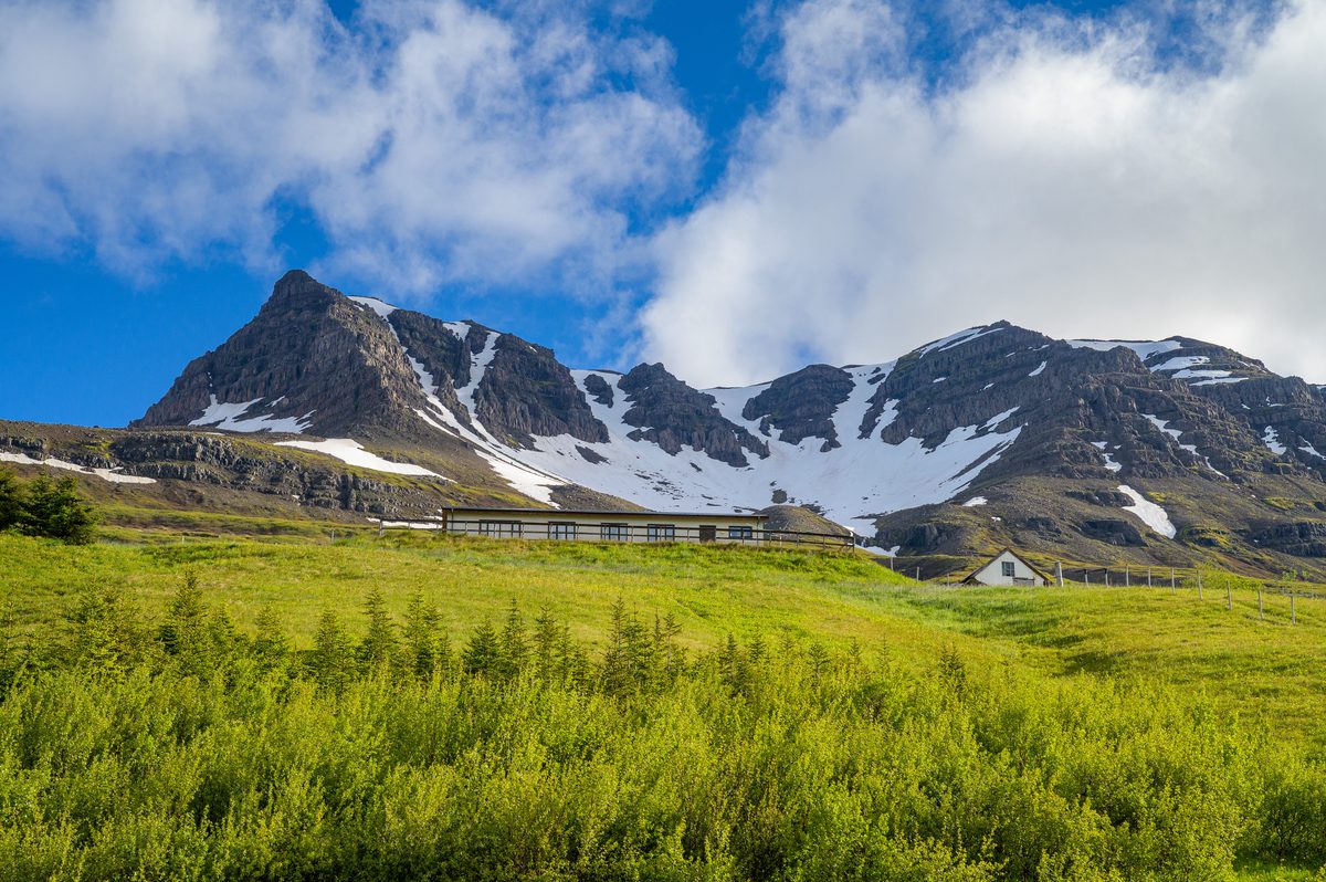 A jagged mountain with lingering snow rises above a luminous green meadow and a few small buildings under broken clouds.