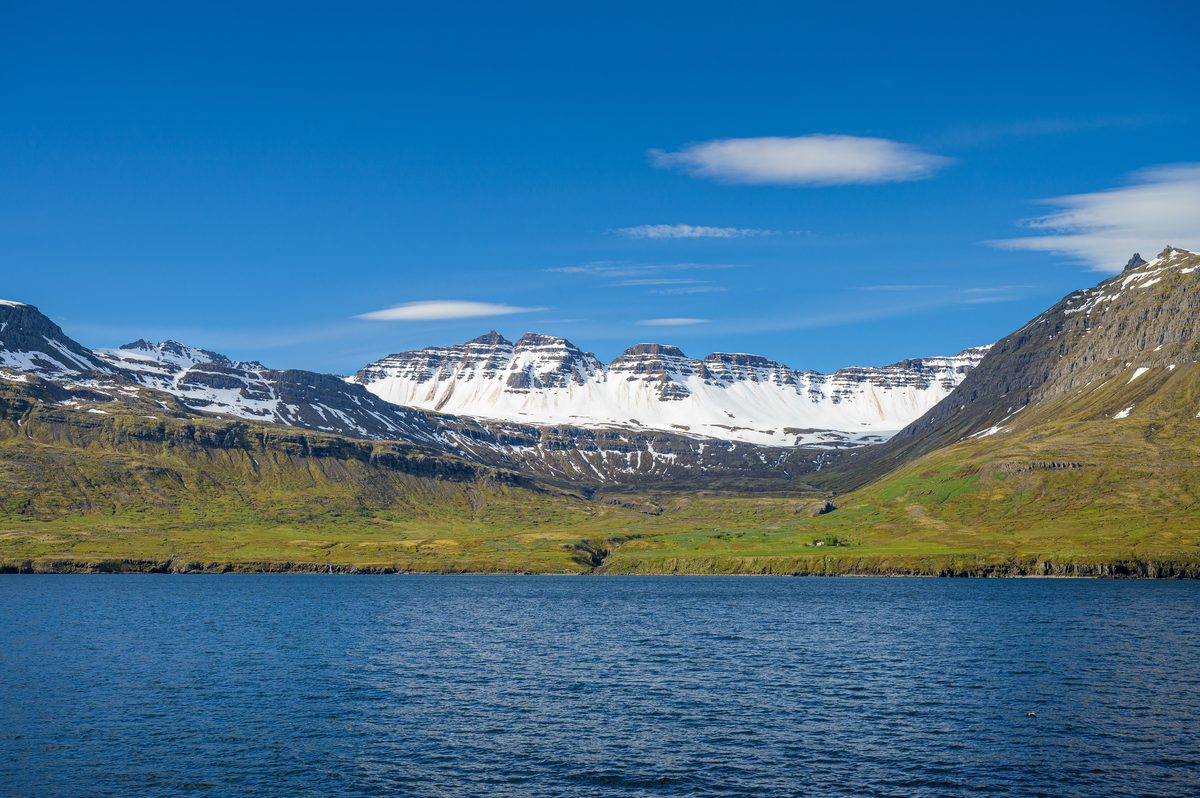 Dark blue fjord water fills the foreground beneath a broad mountain bowl striped with snow and green slopes.