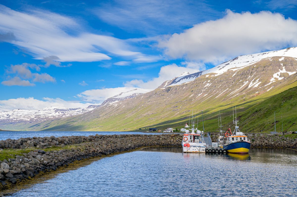 Two colorful fishing boats are moored inside a small rock breakwater beneath the sweeping mountains of Mjóifjörður.