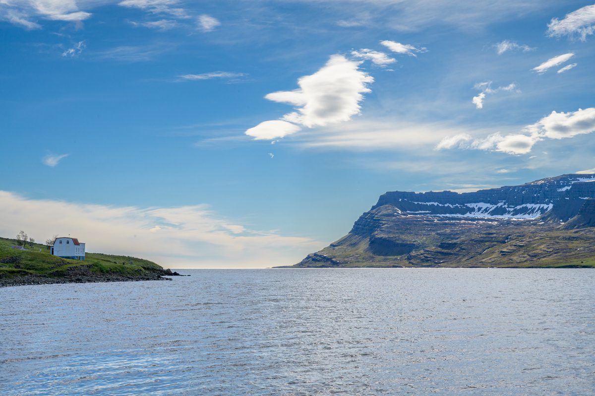 A sculpted white cloud hangs above a flat-topped mountain at the mouth of the fjord, framed by still water and grassy shore.