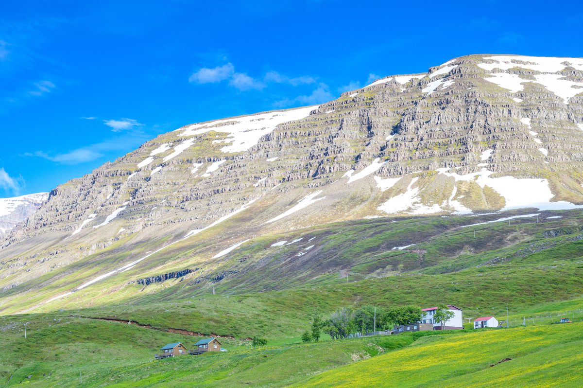 Scattered homes rest on intensely green pasture beneath a massive terraced mountain capped with late snow.