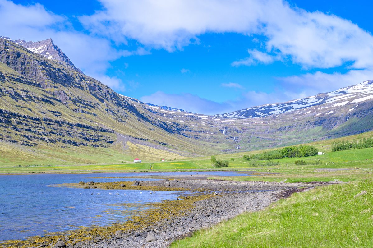 A curving pebbled shoreline and shallow tidal water lead toward a green valley enclosed by steep mountains.