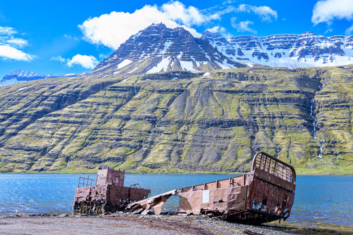 A rusted landing craft lies at the water's edge beneath sharply layered mountains and bright summer clouds.