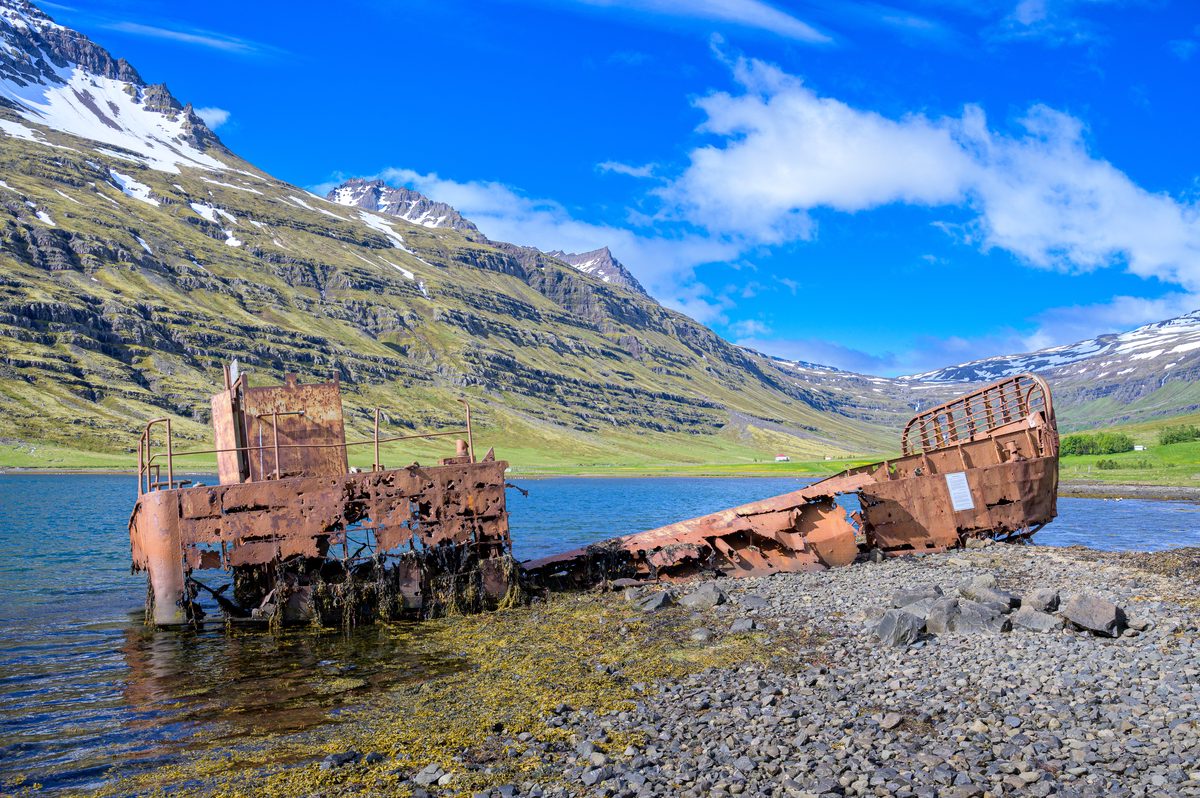 The broken landing craft stretches along the rocky shore, backed by blue water and snow-streaked mountains.