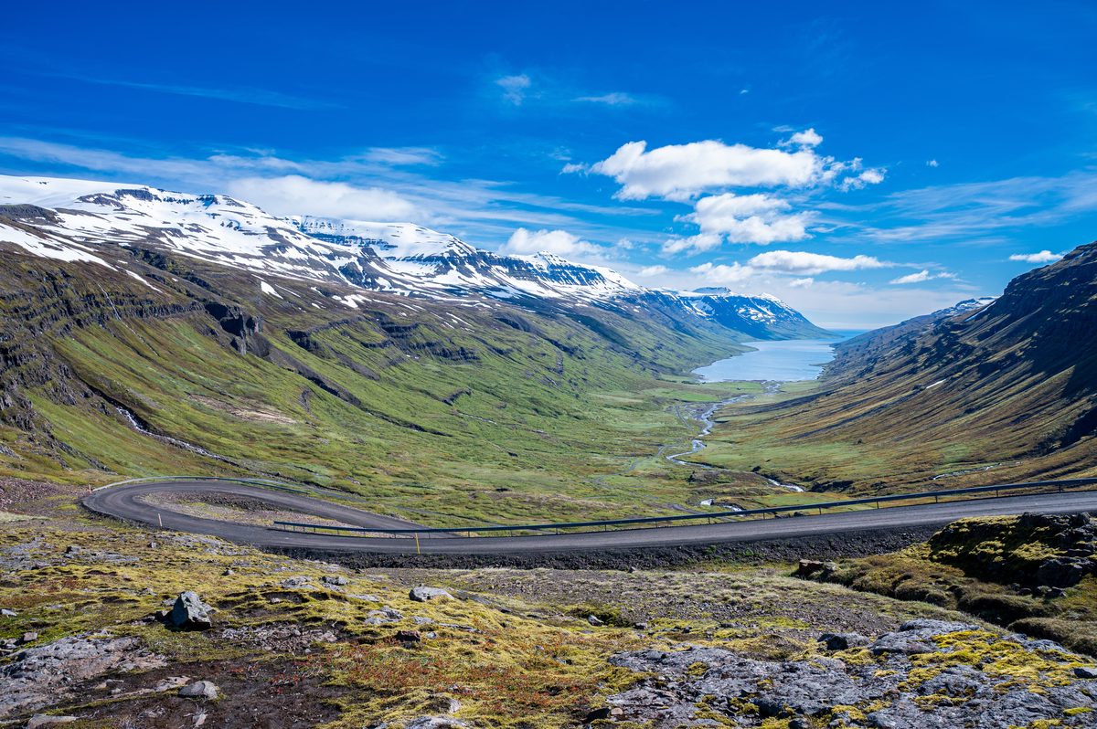 A narrow mountain road curves high above a green valley and distant fjord between ridges patched with snow.