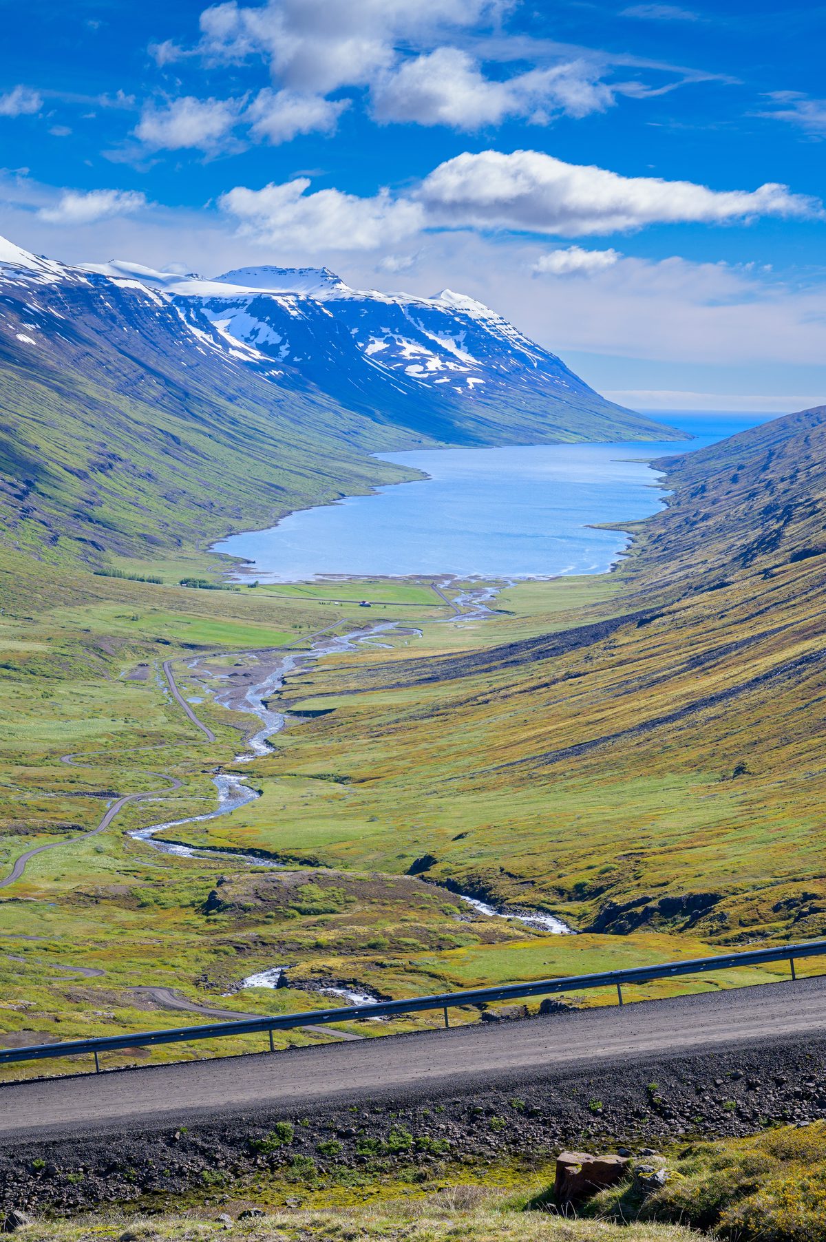 A vertical view looks down a broad green valley toward the blue fjord, with a winding road crossing the foreground.