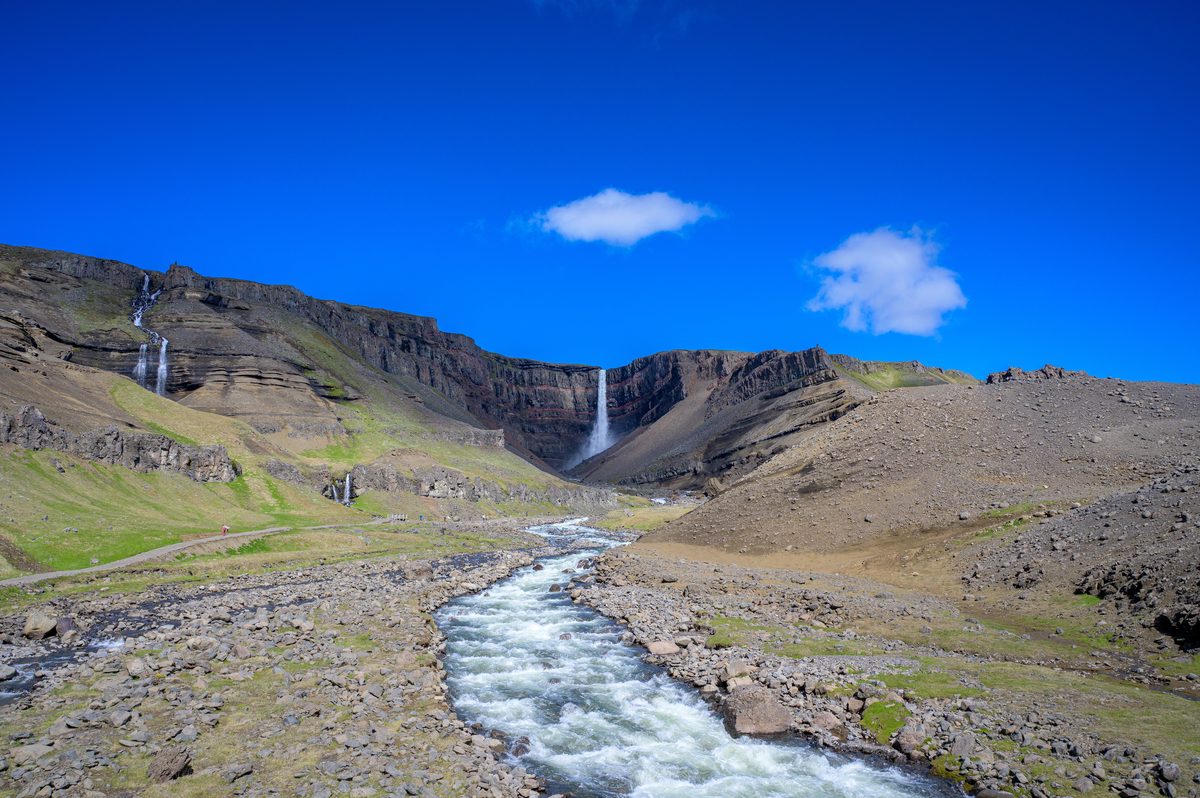 A fast river runs from a tall waterfall in a broad volcanic amphitheater under a clear blue sky.