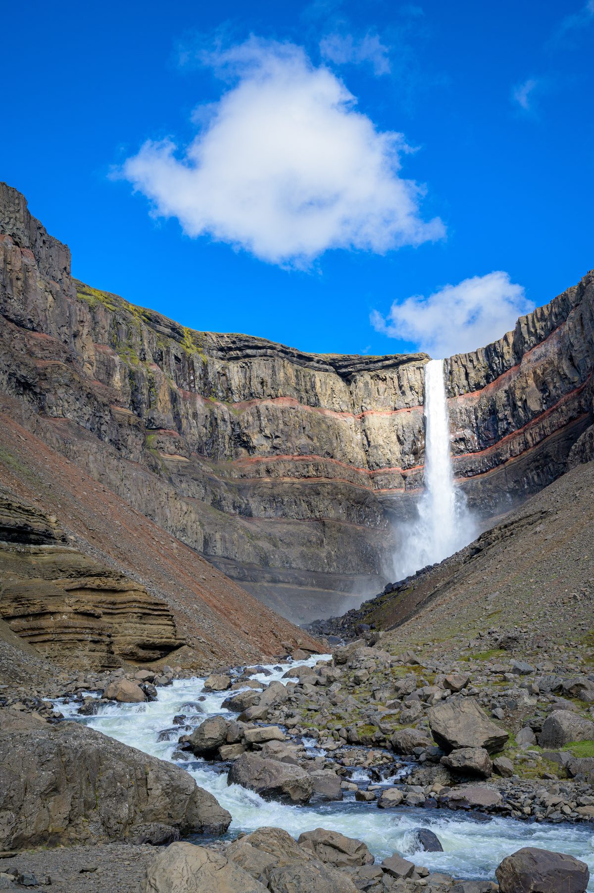 A tall white waterfall drops from a dark layered cliff into a rocky stream beneath a single summer cloud.
