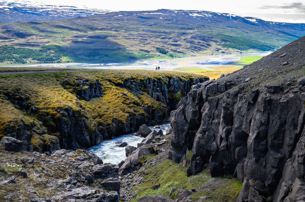 Foaming water twists through a deep basalt gorge while sunlit green lowlands and mountains open beyond.