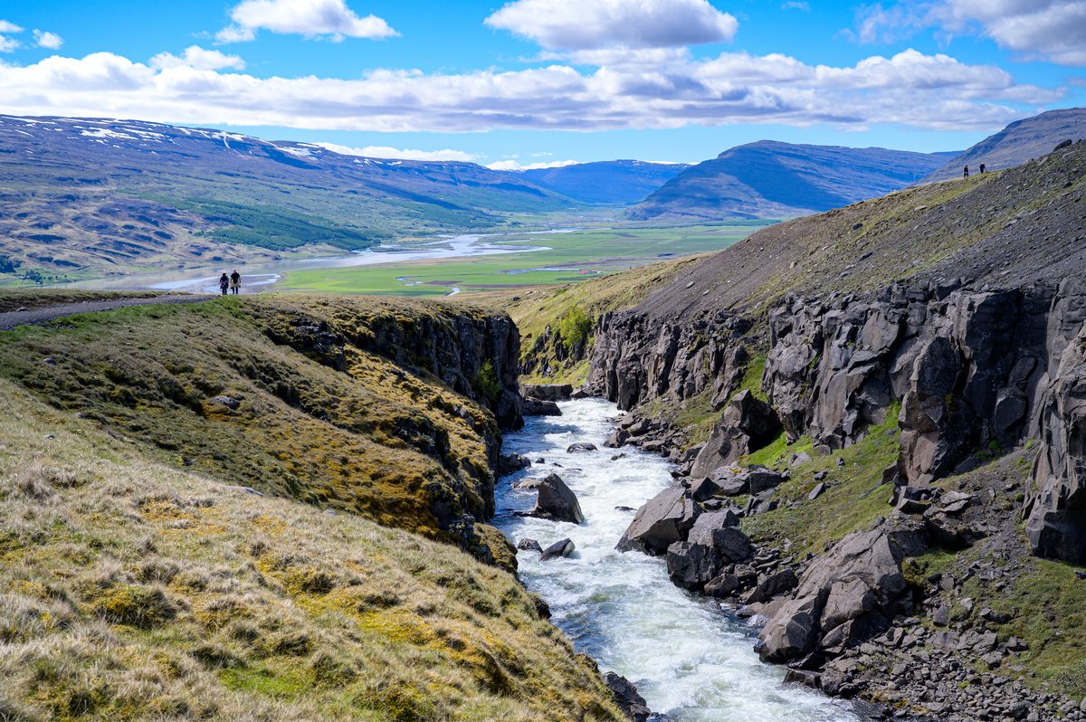 A swift river runs between tall, angular basalt walls with a broad green valley and distant mountains beyond.