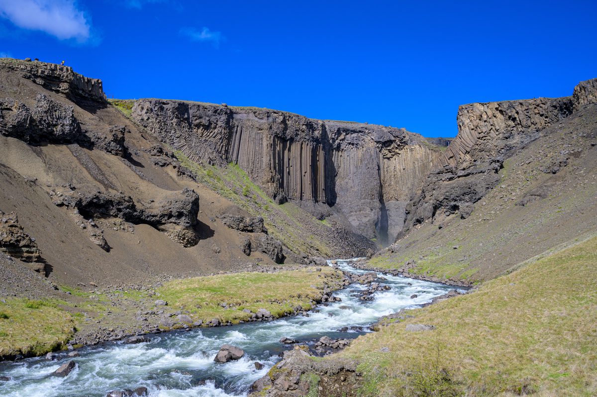 A milky stream curves below a cliff of straight basalt columns in a spare volcanic valley.