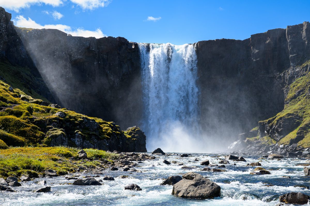 A broad waterfall drops over a dark ledge into a rocky river, edged by bright moss and backlit spray.