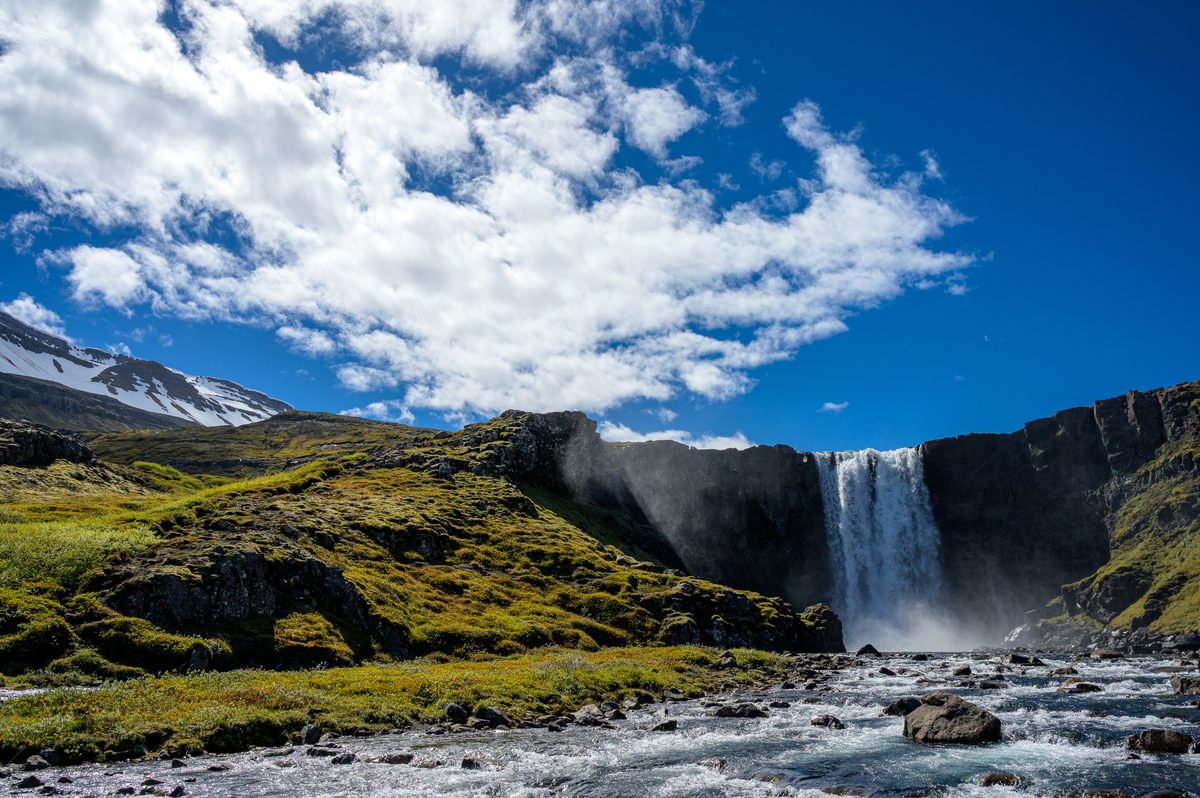 Sunlit clouds billow over a broad waterfall and moss-covered ledges while the river runs through dark foreground rocks.