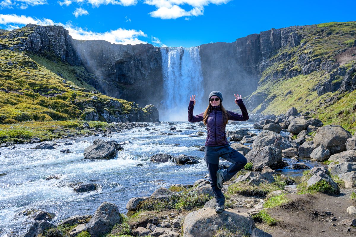A traveler balances on streamside rocks with arms raised in front of a broad waterfall and green basalt cliffs.