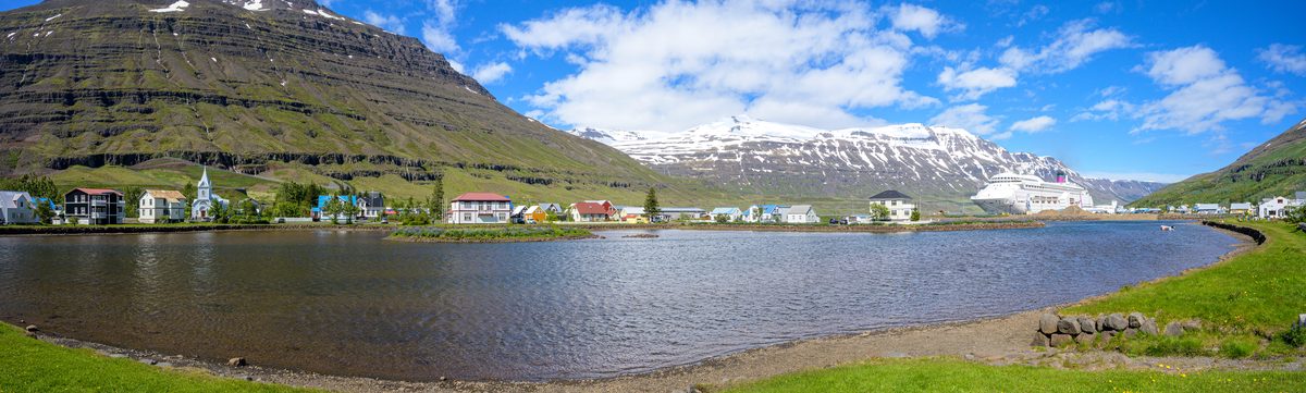 A wide panorama spans the calm lagoon, colorful town, and immense terraced mountains surrounding Seyðisfjörður.