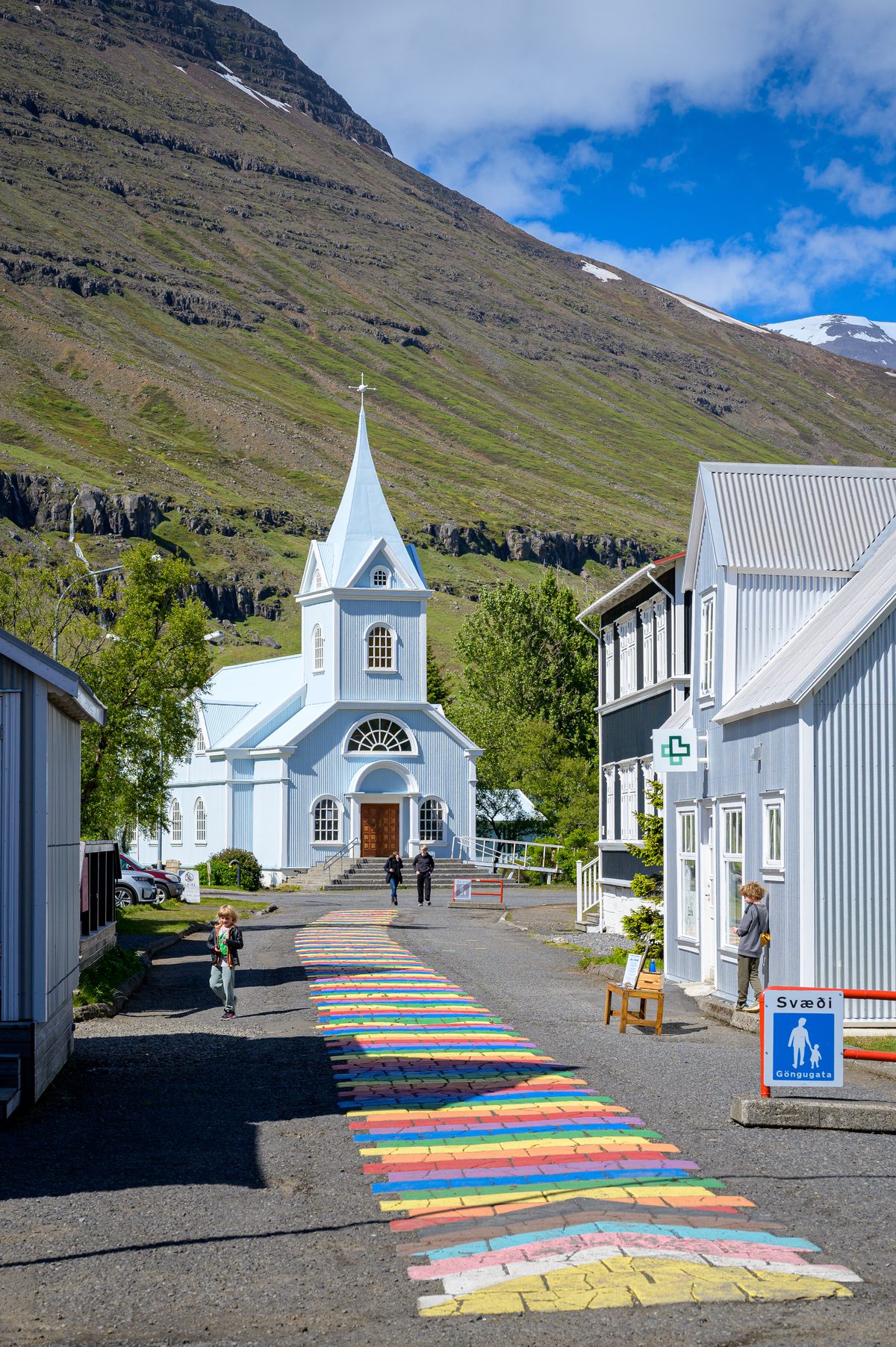 A vertical street view follows the rainbow-painted pavement toward Seyðisfjörður's blue church and green mountains.