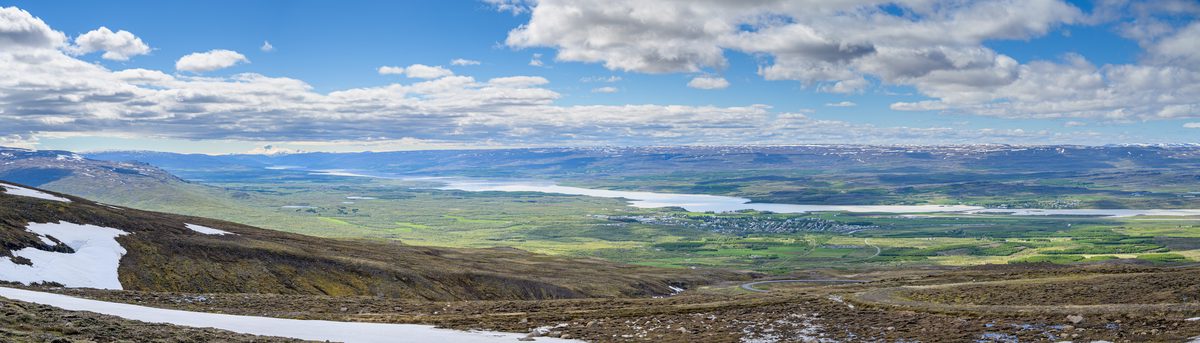 A panoramic view from the snowy mountain pass opens across green lowlands, lakes, and distant blue ridges.