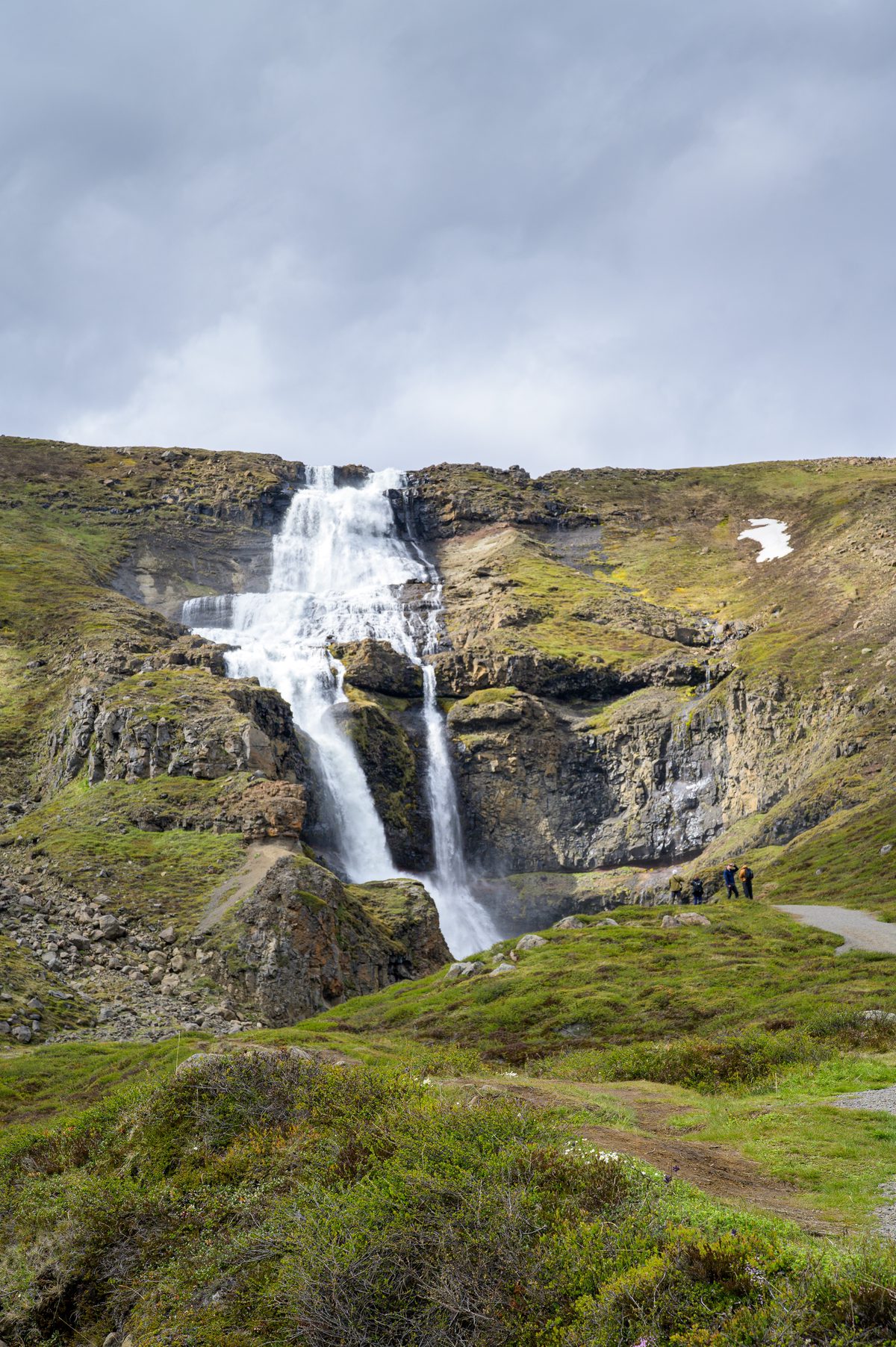 A stepped waterfall streams down a rugged green mountainside with small patches of lingering snow.