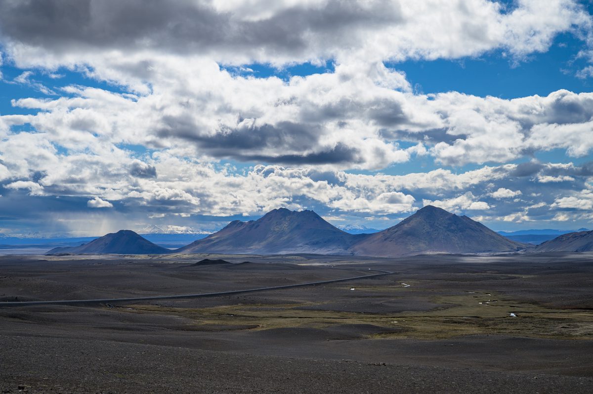 Dark triangular volcanic hills rise from a vast black plain under dramatic layers of sunlit cloud.