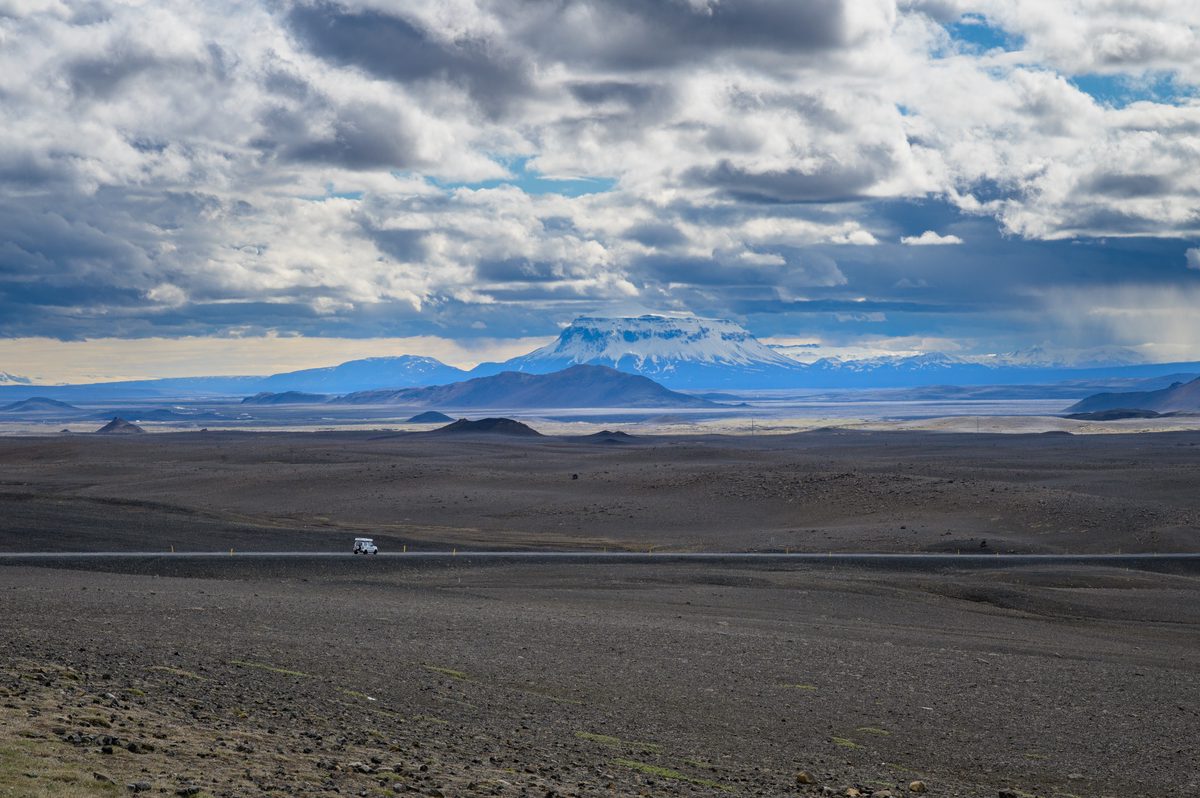 A distant curtain of rain falls beneath broken clouds over a broad charcoal plain and blue mountain horizon.