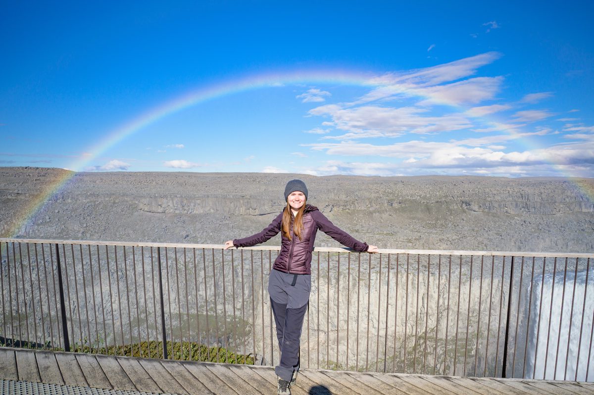 A traveler stands at the viewpoint railing with arms open as a rainbow rises from Dettifoss behind her.