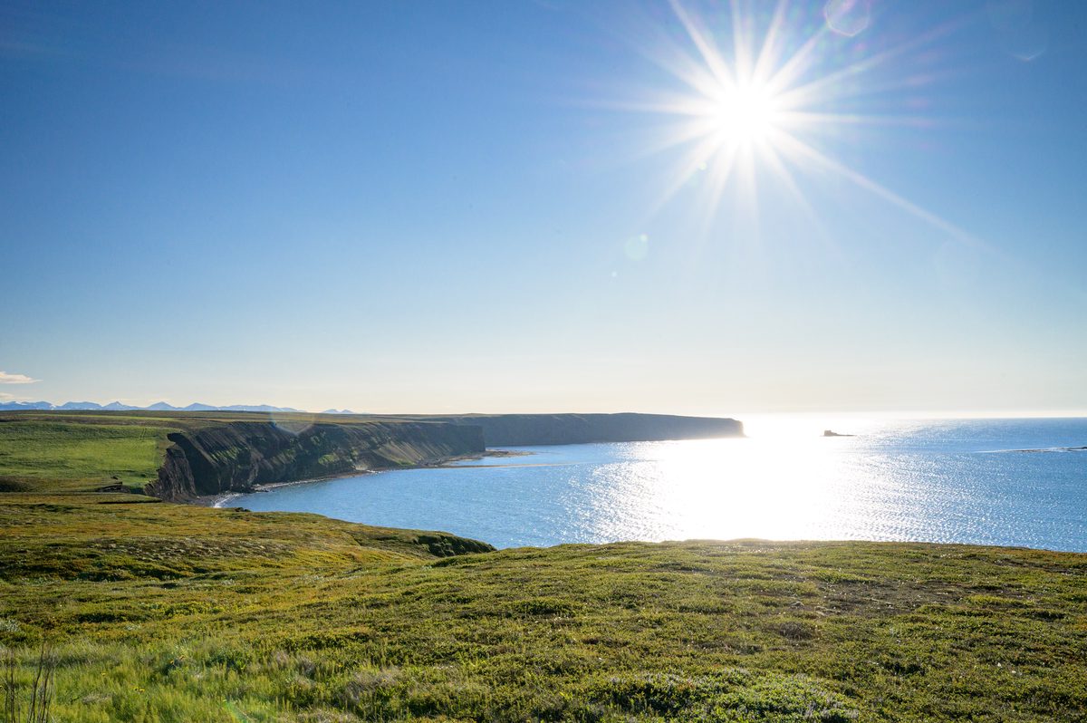 The low sun sparkles across a broad blue bay below green sea cliffs on the Tjörnes peninsula.