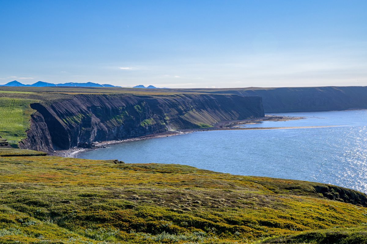 Long green cliffs curve around a blue bay beneath a clear sky, their dark layers dropping sharply to the water.