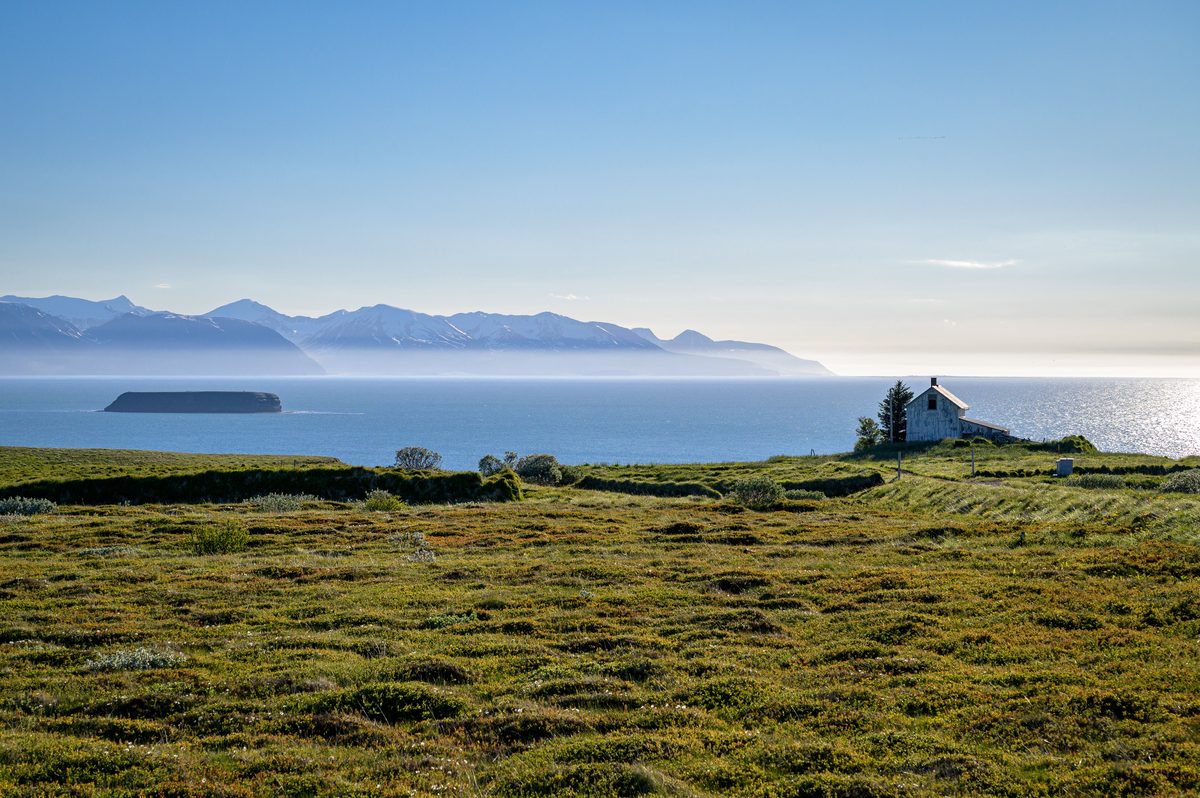 A solitary white building rests in a green coastal meadow above silver-blue water, islands, and distant mountains.