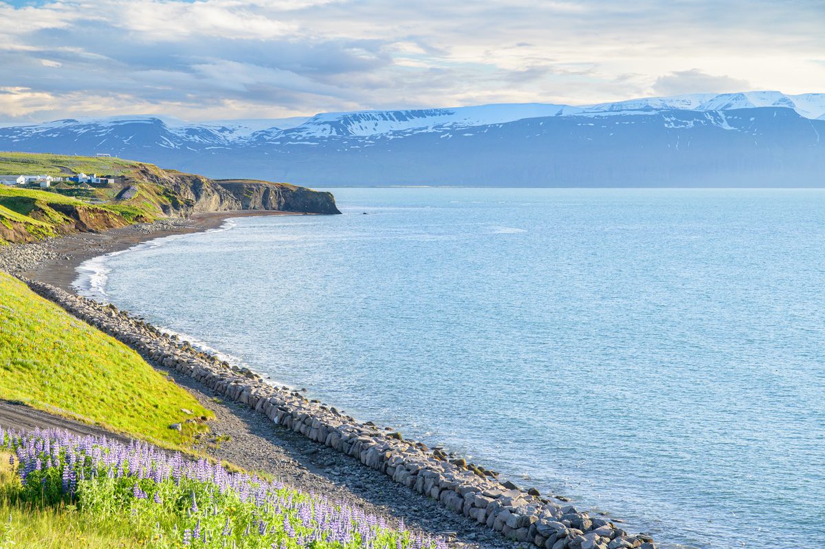 A stone-lined shore curves along clear blue water toward distant snow-striped mountains under soft evening cloud.