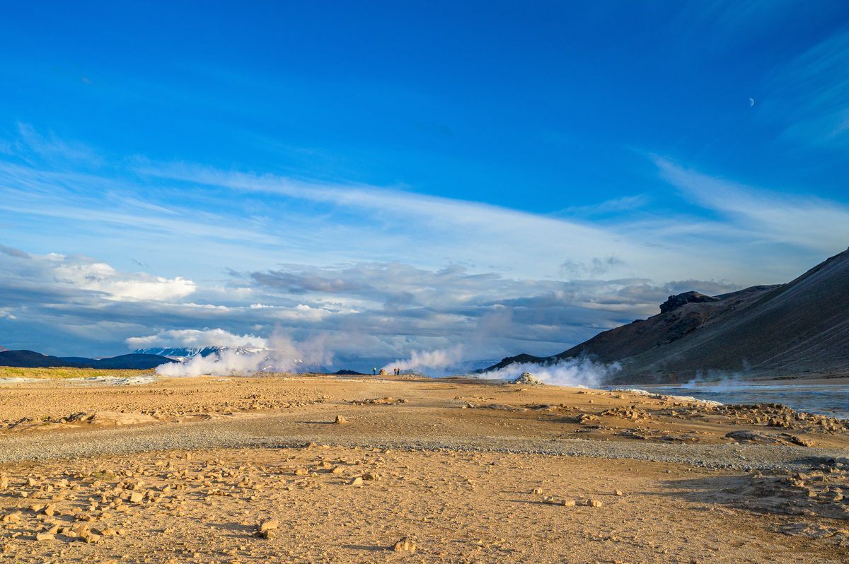 White geothermal steam drifts across a pale ochre plain beneath a sweeping blue sky at Hverir.
