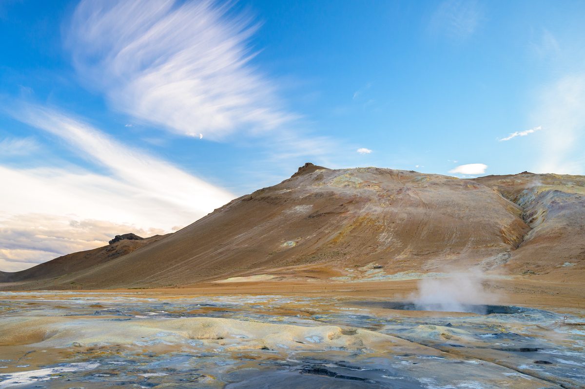 A rust-colored volcanic ridge rises above blue-gray mineral ground and wisps of steam under streaked clouds.