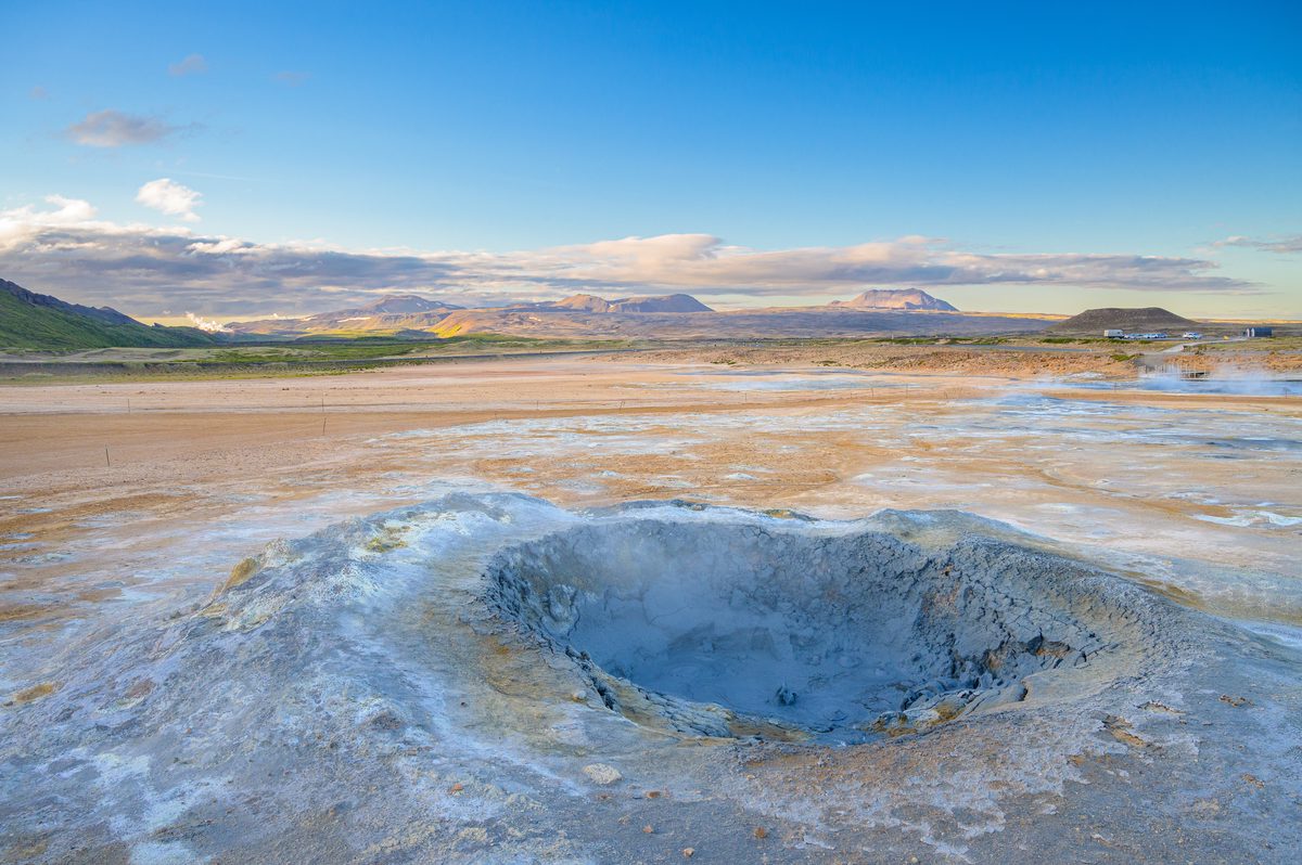 A blue-gray mud pot bubbles inside a small circular basin surrounded by white, rust, and ochre mineral deposits.