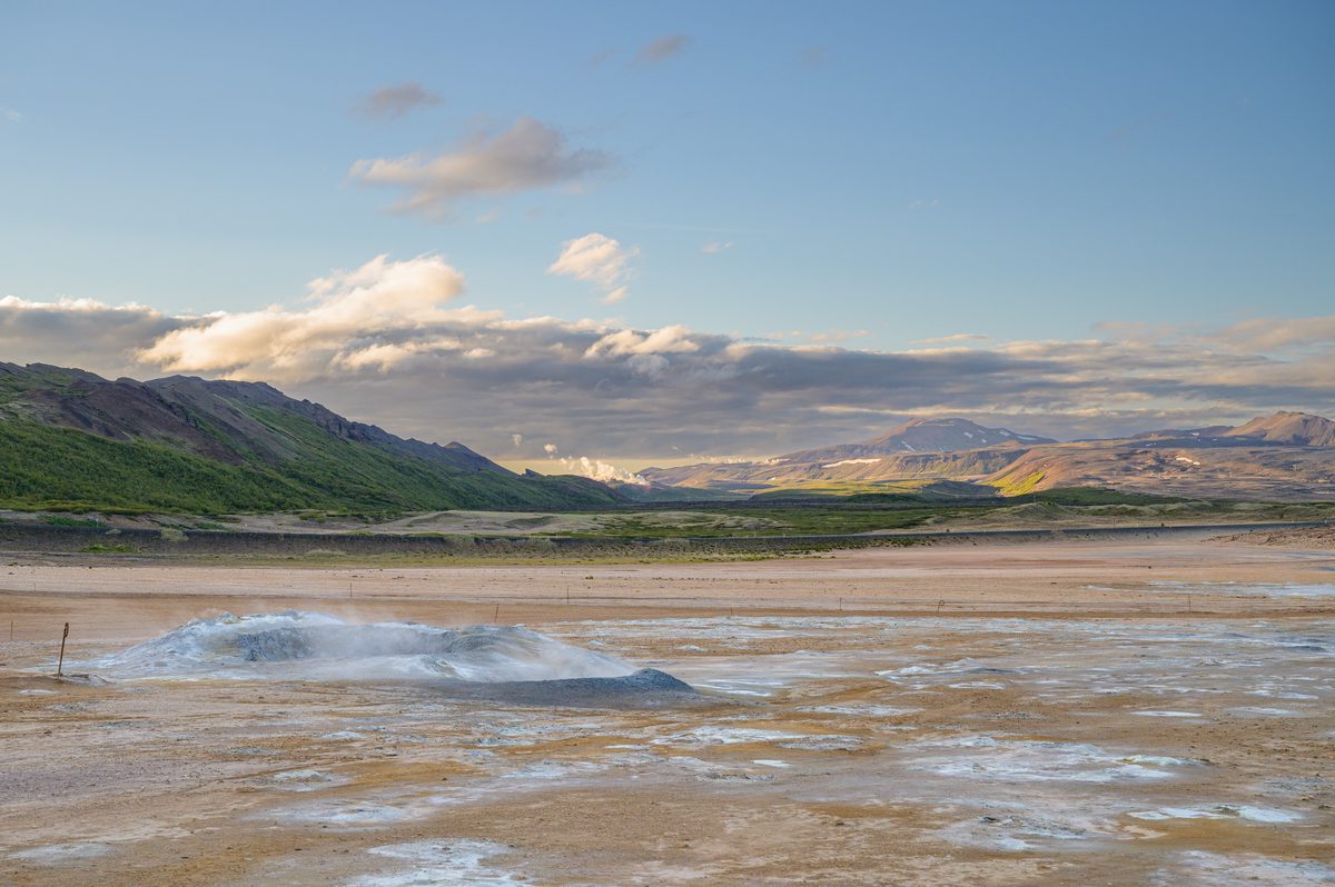Thin steam trails across a broad blue-gray and ochre geothermal plain with green mountains in the distance.