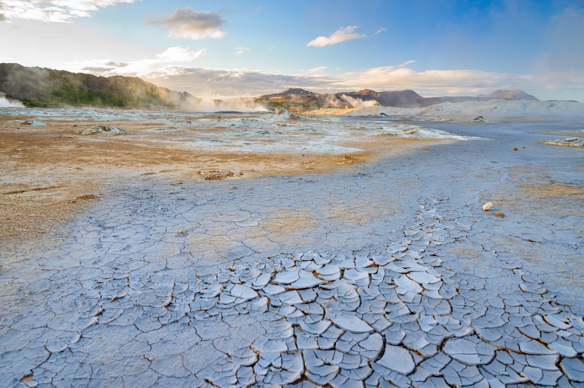 Polygonal cracked clay fills the foreground while steam rises from pale mineral ground under dramatic evening clouds.