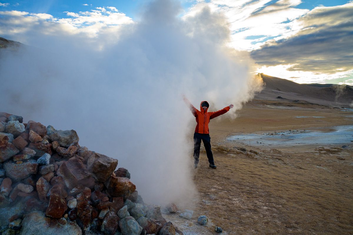 A traveler in a red jacket stands with arms open beside a powerful white geothermal steam vent.
