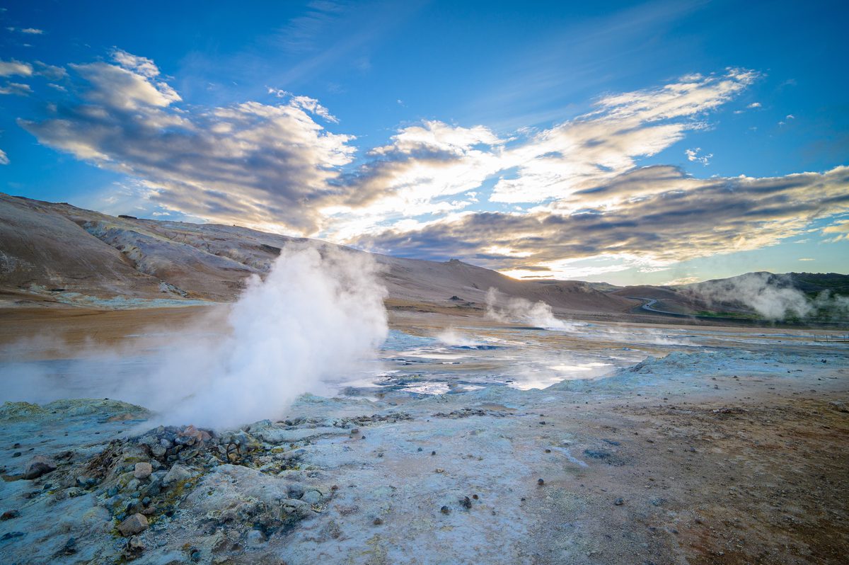 A tall steam plume glows against the low sun and textured clouds above the blue-gray geothermal field.