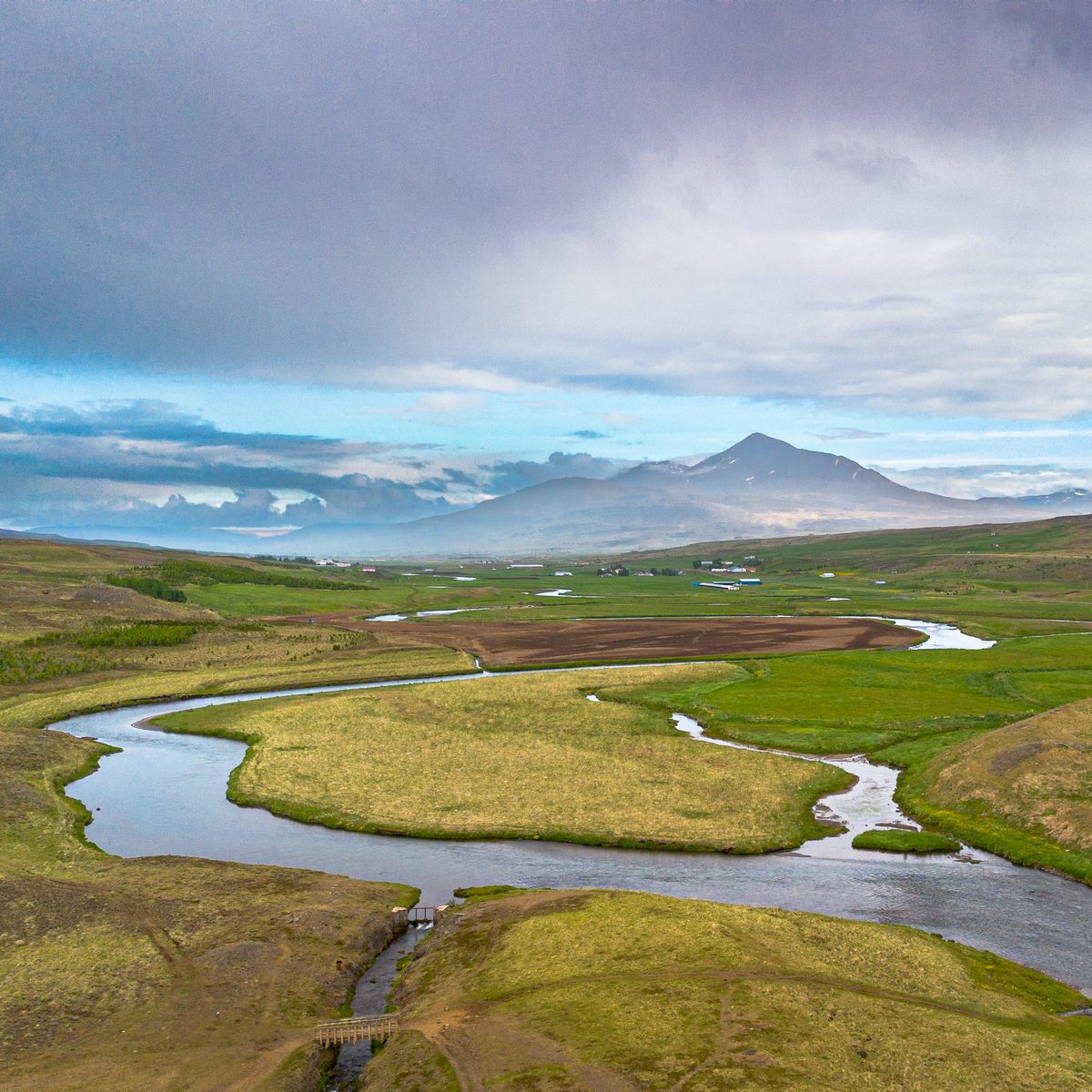 A square aerial view follows a winding river around bright green islands and low banks with a conical mountain in the distance.