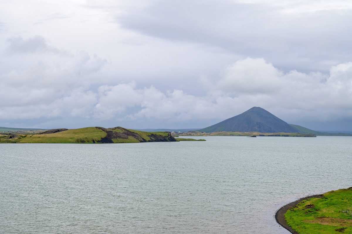 Green pseudocraters rise from the pale water of Lake Mývatn beneath misty mountains and rain-filled clouds.