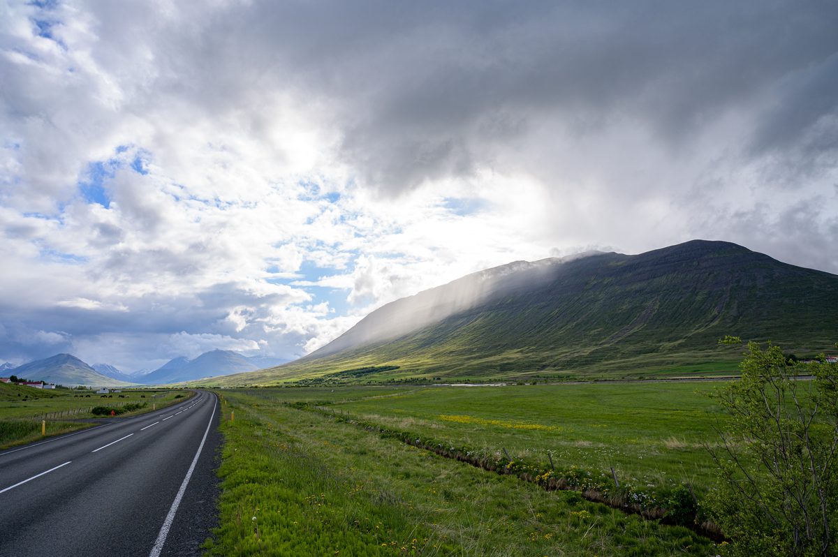 The Ring Road runs straight toward a green mountain whose upper slopes disappear into streaming gray cloud.