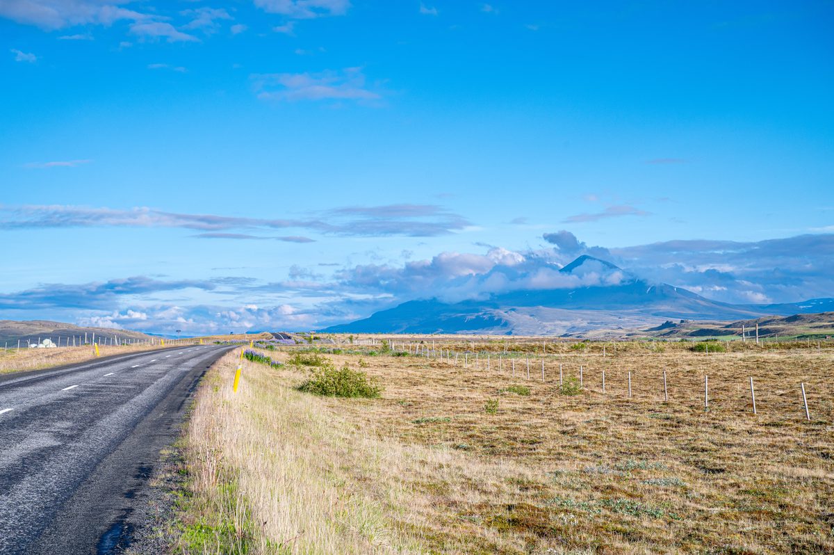 A quiet paved road crosses pale open country toward distant snow-striped mountains under a bright blue sky.