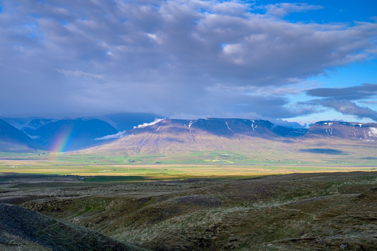 A faint rainbow touches a broad green valley below layered mountains and dramatic evening clouds.